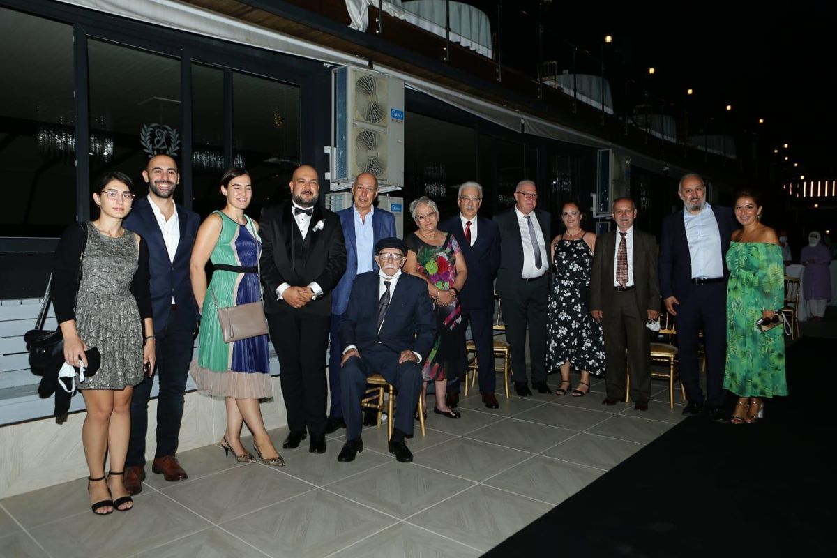 Bayoğlu ailesinden on üç kişi, akşam saatlerinde bir mekânın önünde bir arada poz veriyor; ortada sandalyede oturan yaşlı bir adam var.