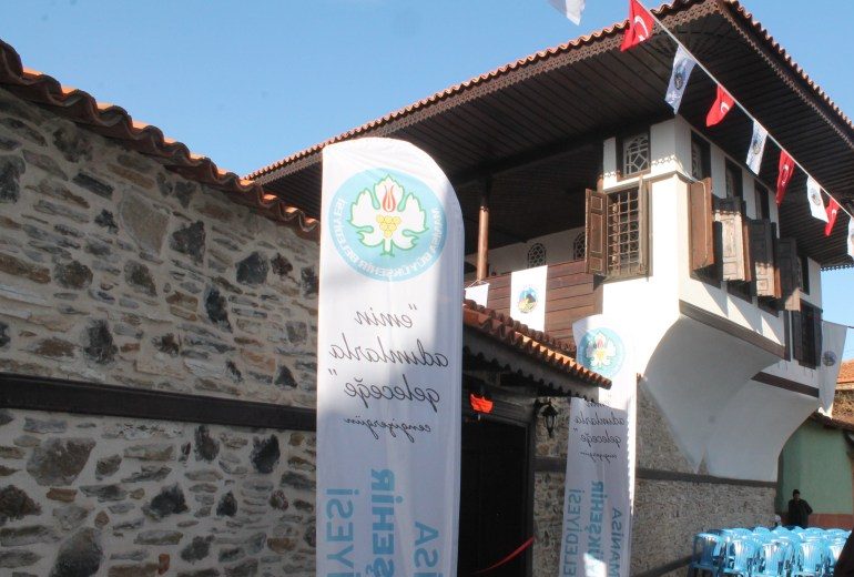 Taş duvarlı sokaktan görünen, ahşap çıkmalı ve geniş saçaklı iki katlı Beyler Evi; sokakta bayraklar ve açılış pankartları var.