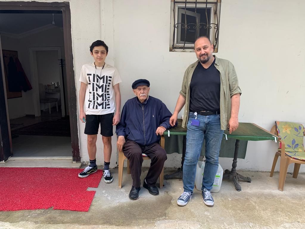 Furkan Bayoğlu (sağda, ayakta), babası Mehmet İlhan Bayoğlu (ortada, sandalyede) ve torun Mehmet İlhan (solda), bir evin kapısı önünde.